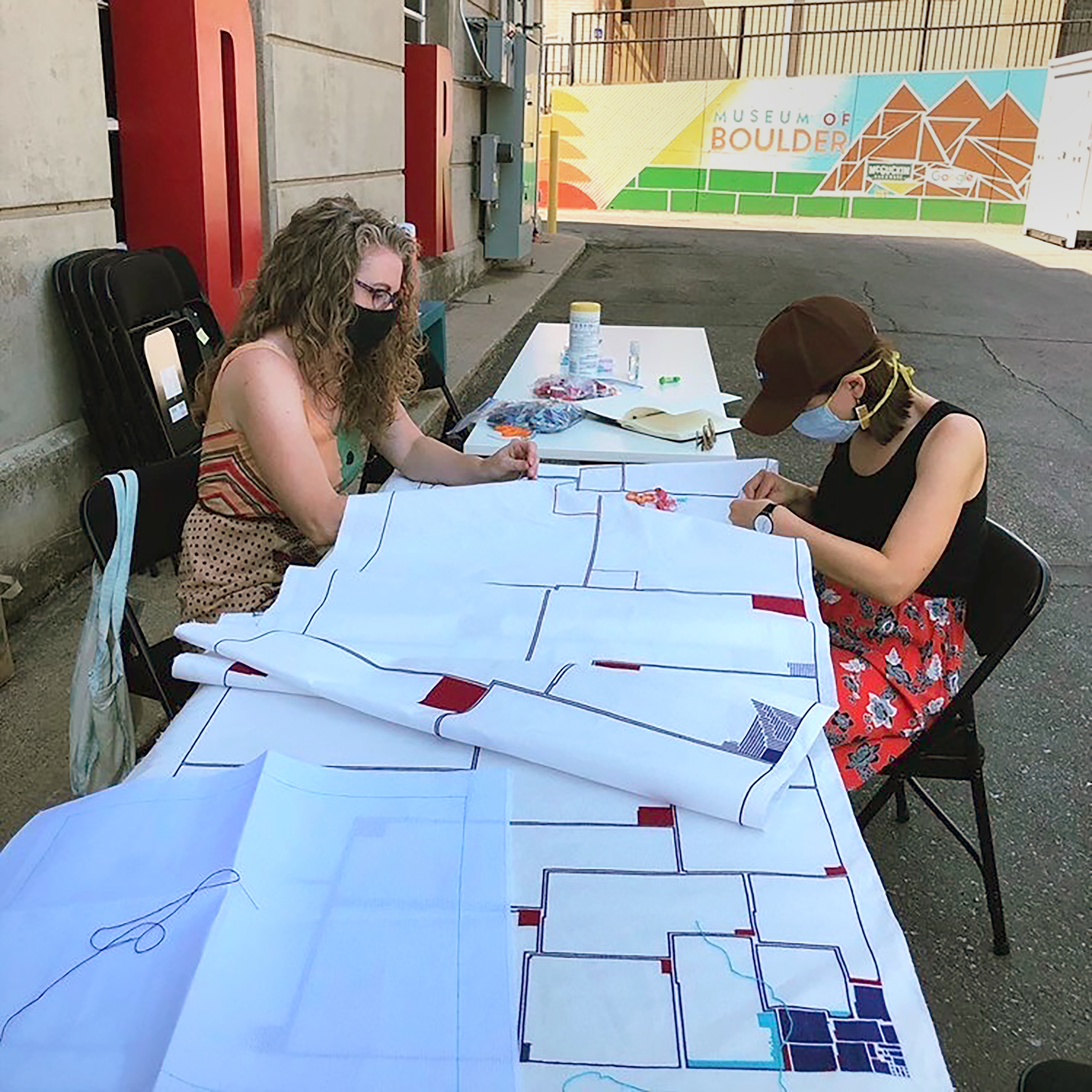 two people with masks on at a table, sewing